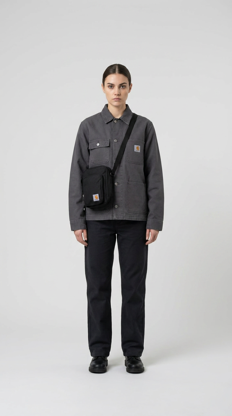 Kvinde i grå Carhartt-jakke, sorte bukser og sort skuldertaske foran en hvid baggrund.