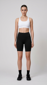 Kvinde i hvid sports-bh og sorte cykelshorts stående mod en lys grå baggrund.