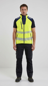Mand iført gul refleksvest med refleksstriber og sorte arbejdsbukser på en lys baggrund.
