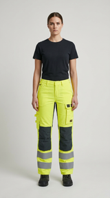 Kvinde i sort t-shirt og neongule arbejdsbukser med reflekser og knæpuder foran en grå baggrund.
