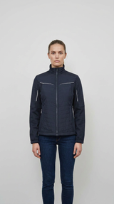 Kvinde i en mørkeblå quiltet jakke med refleksdetaljer og blå jeans mod en enkel baggrund.