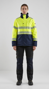 Kvinde i gul og blå high-vis arbejdsjakke med reflekser og mørke arbejdsbukser mod en grå baggrund.