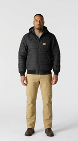 Mand i sort quiltet Carhartt-jakke, beige bukser og brune støvler mod en hvid baggrund.