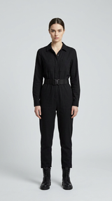 Kvinde i sort langærmet jumpsuit med bælte og sorte støvler mod en grå baggrund.