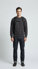 Mand i mørkegrå sweatshirt og mørkeblå jeans med en sort rygsæk foran en hvid baggrund.