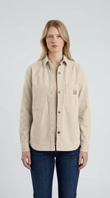 Kvinde med lyst hår iført en beige Carhartt-overskjorte og blå jeans mod en hvid baggrund.