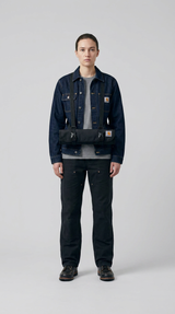 Person i mørk denimjakke og sorte arbejdsbukser med en sort Carhartt-værktøjsrulle over brystet.