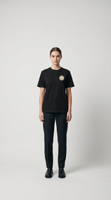 Kvinde i sort t-shirt med logo, sorte bukser og støvler mod en hvid baggrund.