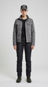 Kvinde iført grå jakke, mørke jeans og kasket stående mod en lys baggrund.