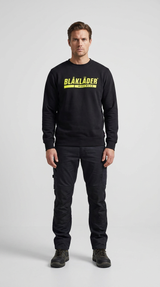Mand i sort Blåkläder sweatshirt og sorte arbejdsbukser mod en grå baggrund.