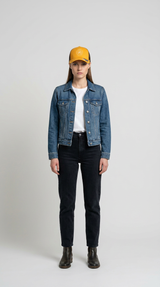 Kvinde i blå denimjakke, gul kasket og sorte jeans stående mod en grå baggrund.