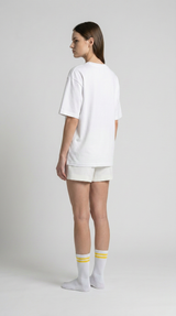 Kvinde i hvid t-shirt, shorts og hvide strømper med gule striber set bagfra mod en lys baggrund.