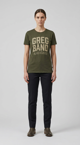 Kvinde i grøn t-shirt med "GREG BÅND" tekst, mørke bukser og støvler mod en grå baggrund.