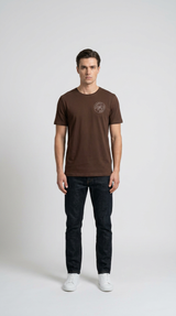 Mand i brun t-shirt med lille logo, mørkeblå jeans og hvide sneakers mod en lys baggrund.