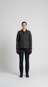 Kvinde iført en koksgrå arbejdsjakke, mørke jeans og brune handsker mod en enkel hvid baggrund.