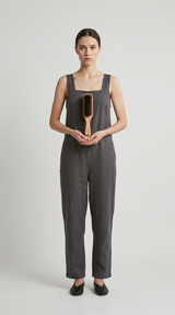 Kvinde i mørkegrå hør-jumpsuit holder en hårbørste af træ mod en neutral baggrund.
