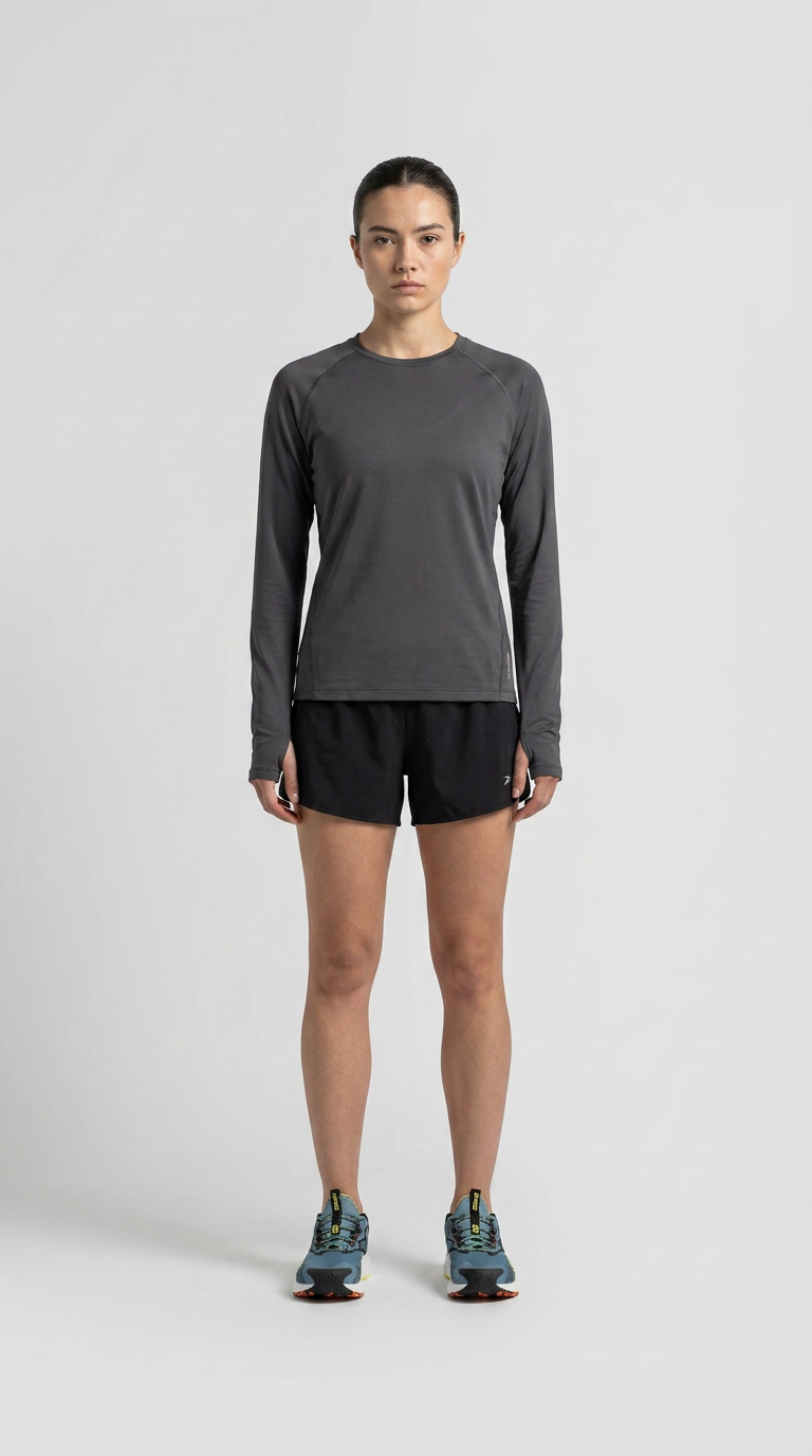 Kvinde i grå langærmet træningstrøje, sorte shorts og blå løbesko mod en grå baggrund.