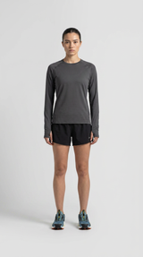 Kvinde i grå langærmet træningstrøje, sorte shorts og blå løbesko mod en grå baggrund.