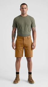 Mand med tatoveringer iført grøn t-shirt, brune arbejdsshorts og støvler foran en hvid baggrund.