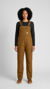 Kvinde med krøllet hår iført brune Carhartt-overalls og en sort langærmet trøje mod en grå baggrund.