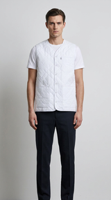 Mand i hvid quiltet vest, hvid t-shirt og mørke bukser mod en lys baggrund.