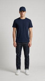 Mand i mørkeblå t-shirt, mørke jeans, kasket og hvide sneakers mod en lys grå baggrund.