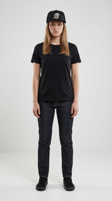Kvinde i sort t-shirt, mørke jeans og sort kasket står foran en lys, ensfarvet baggrund.