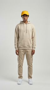 Ung mand i beige hættetrøje, cargobukser, gul kasket og hvide sneakers mod en grå baggrund.