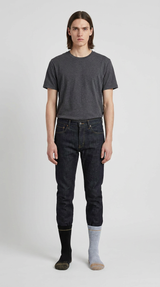 Mand i grå t-shirt, mørkeblå jeans og umage sorte og grå sokker foran en hvid baggrund.