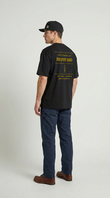 Mand i sort T-shirt med 'Tattoos by Kasper Baum' tekst på ryggen, blå bukser og en sort kasket.