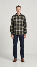 Mand i grøn- og sortternet flannelsskjorte, mørkeblå jeans og brune støvler mod en grå baggrund.