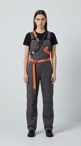 Kvinde i grå Carhartt-overalls med orange remme og en sort t-shirt mod en lys baggrund.