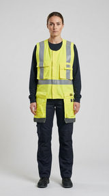 Kvinde i gul high-visibility vest og mørke arbejdsbukser mod en lys grå baggrund.