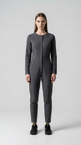 Kvinde i en langærmet grå jumpsuit og sorte sneakers mod en lys grå baggrund.