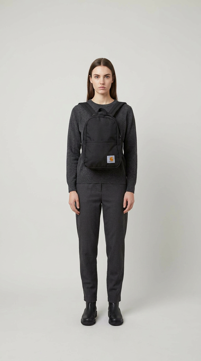 Kvinde iført gråt tøj med en sort Carhartt-rygsæk på brystet mod en lys baggrund.
