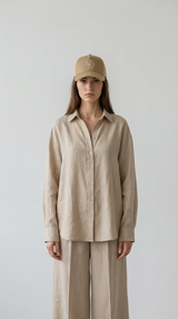 Kvinde i beige hørskjorte, matchende bukser og en brun kasket mod en enkel grå baggrund.