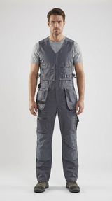 Mand iført grå ærmeløse arbejds-overalls med mange lommer over en t-shirt mod en lys baggrund.