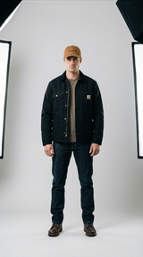 Mand iført sort Carhartt-jakke, brun kasket og mørkeblå jeans i et lyst fotostudie.